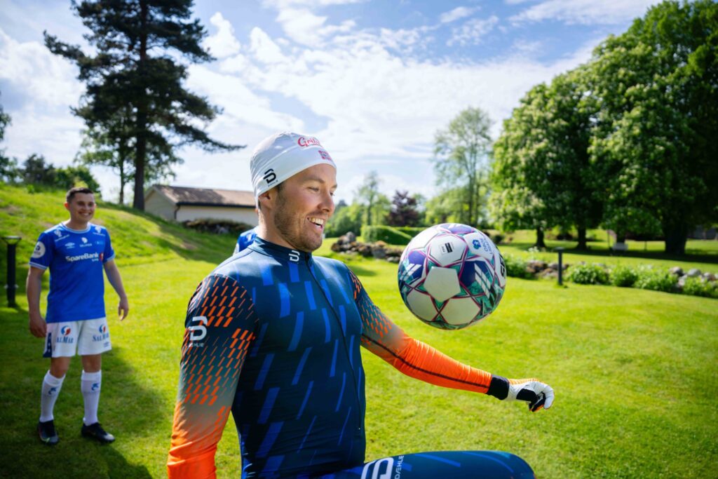 Mann i treningstøy jonglerer med en fotball på en gressplen mens en fotballspiller ser på